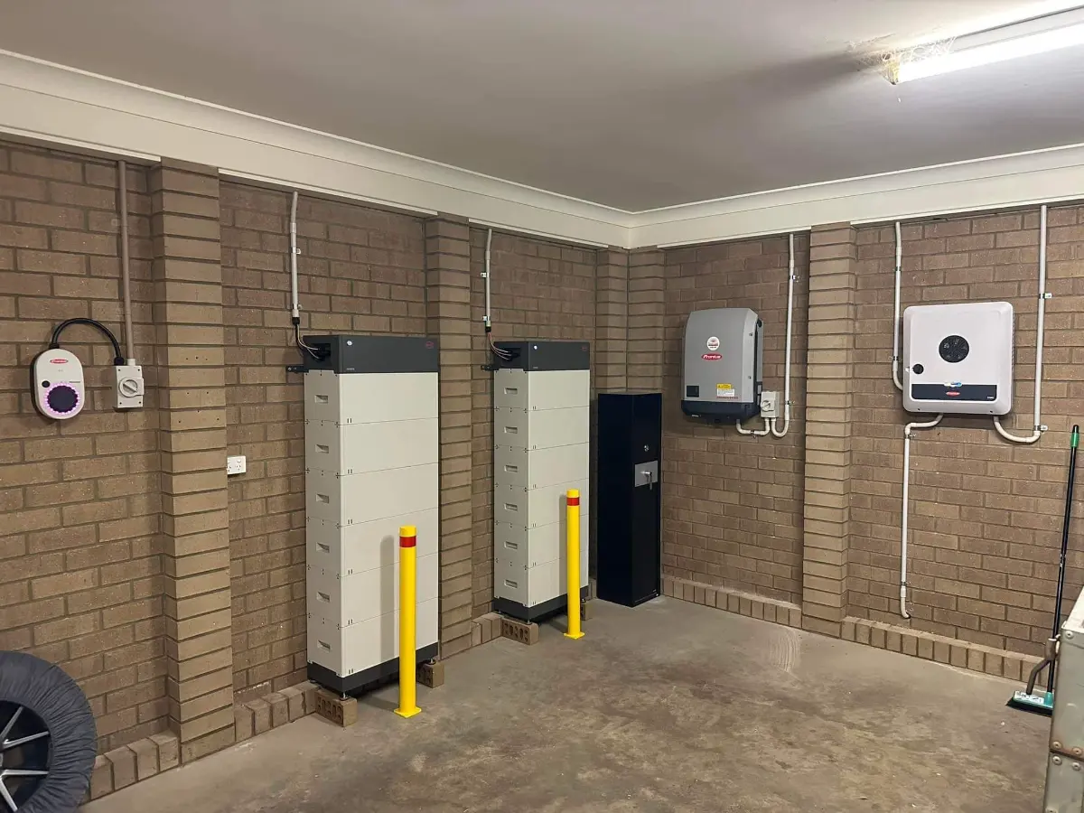 Garage with two white battery towers, two Fronius inverters and an EV charger mounted on brick walls, with yellow bollards